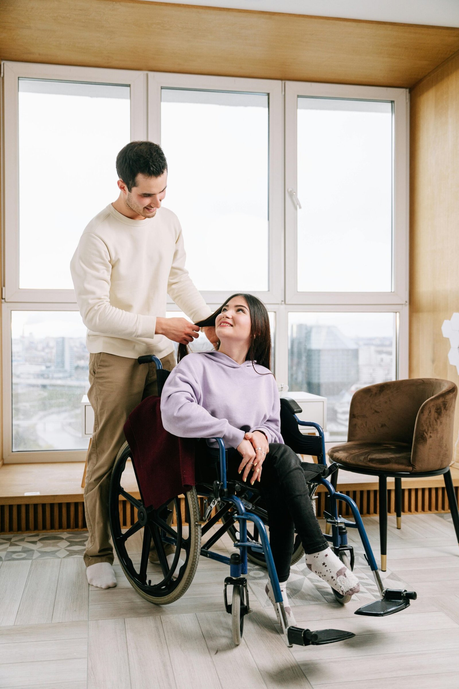 A loving couple shares a moment, one sitting in a wheelchair, in a sunlit room.